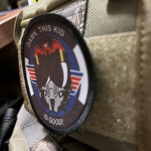 May include: A beige fabric bag with a patch that reads "DAMN THIS KID IS GOOD!" The patch features a cartoon pilot wearing a helmet and goggles.