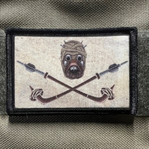 May include: A black and white embroidered patch with a beige background. The patch features a skull with a wrapped head and crossed spears.