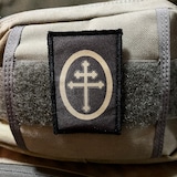 May include: A black patch with a white cross inside a circle. The cross has four arms and a rounded top. The circle has a white outline.