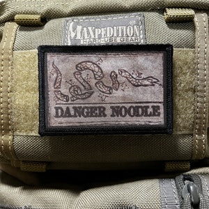 May include: A brown and black patch with a snake design and the text "DANGER NOODLE" on a green Maxpedition bag.
