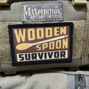 Peut inclure: Un écusson noir et marron avec le texte "Wooden Spoon Survivor" et un dessin de cuillère en bois marron. L'écusson est fixé à un sac en tissu vert et gris avec le texte "Maxpedition Hard-Use Gear" sur une étiquette noire.