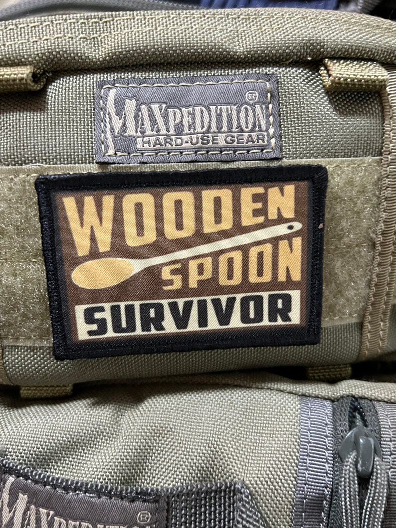 May include: A black and brown patch with the text "Wooden Spoon Survivor" and a brown wooden spoon graphic. The patch is attached to a green and gray fabric bag with the text "Maxpedition Hard-Use Gear" on a black tag.