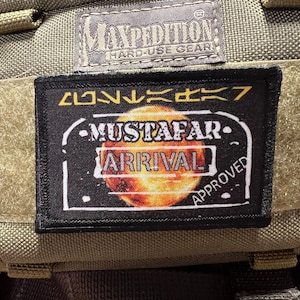May include: A black and white patch with a red and orange planet in the center. The patch has the text "MUSTAFAR ARRIVAL" and "APPROVED" on it. The patch is attached to a green tactical bag.
