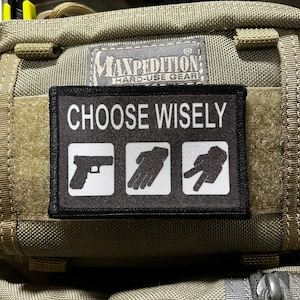 May include: A black patch with white text that reads "Choose Wisely" and three white icons: a gun, a hand with a fist, and a hand with two fingers extended. The patch is attached to a green tactical bag with the Maxpedition logo.