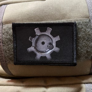Puede incluir: Un parche negro con un diseño blanco en forma de engranaje que presenta un círculo en el centro. El parche está unido a un fondo de tela beige.