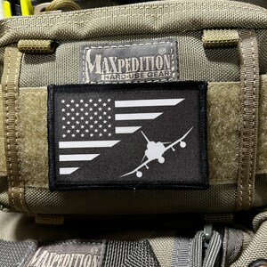 May include: A black and white American flag patch with a white silhouette of a fighter jet on a green tactical bag with the text "Maxpedition Hard-Use Gear" on a black patch.