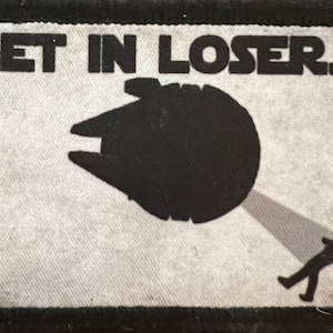 Könnte beinhalten: Ein rechteckiger Aufnäher mit dem Text "GET IN LOSER..." über einer schwarzen Silhouette eines Raumschiffs und einer Person in einem Scheinwerfer. Der Aufnäher hat einen schwarzen Rand und einen hellgrauen Hintergrund.
