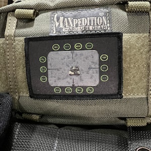 Op de afbeelding: Een groene Maxpedition Hard-Use Gear rugzak met een zwarte patch waarop "DAY, NVG, MPO, IR, RED" staat en die een groene cirkel met een kruisdraad in het midden heeft. De patch is bevestigd aan de voorkant van de rugzak.
