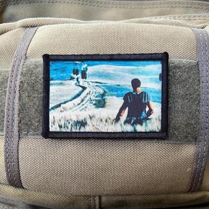 May include: A black and white patch featuring a photograph of a person sitting on a path in a field. The path leads to a woodland in the distance. The sky is blue and cloudy.
