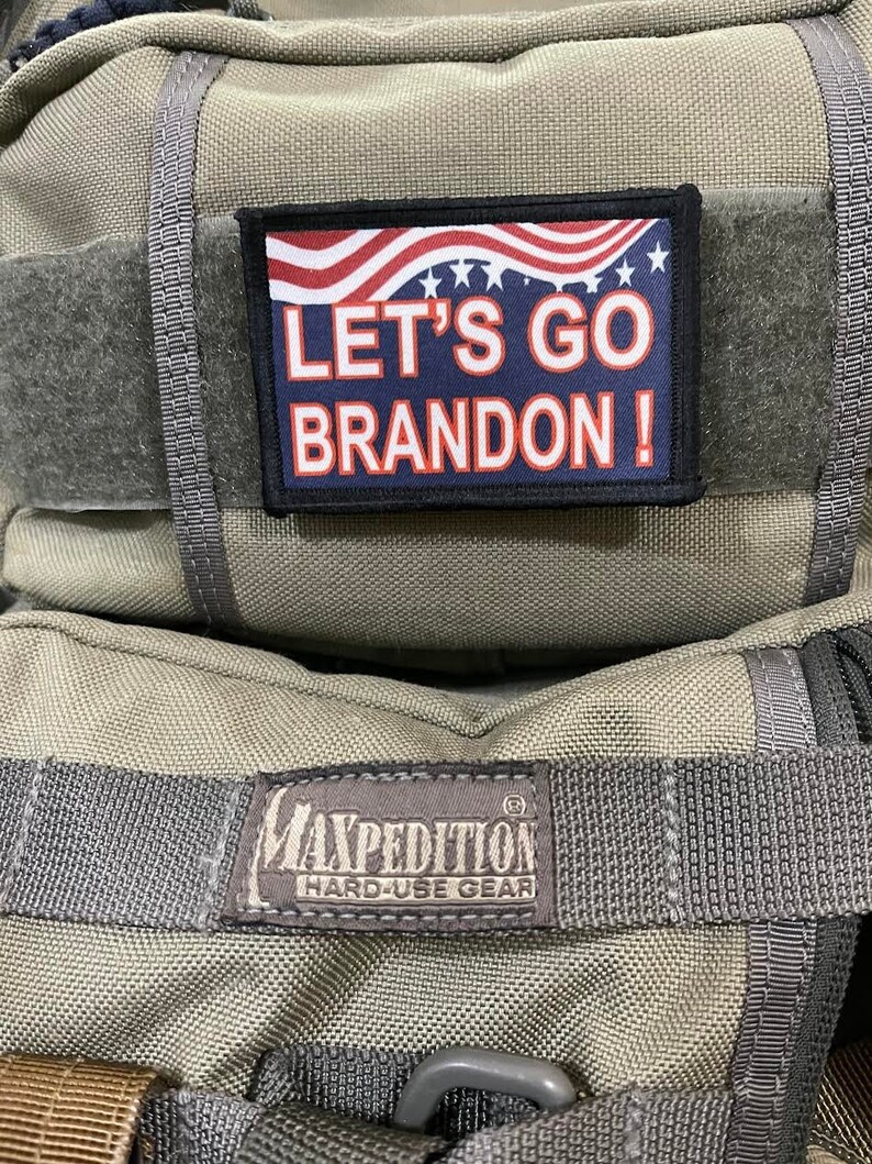 May include: A green Maxpedition backpack with a black patch that says "Let's Go Brandon!" with an American flag design. The backpack has a black strap and a black label that says "Maxpedition Hard-Use Gear".