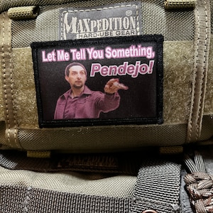Pode incluir: Um remendo preto e cinza com o texto "Let Me Tell You Something, Pendejo!" em letras cor-de-rosa. O remendo apresenta um homem com barba e uma camisa preta, apontando para a direita. O remendo está preso a uma bolsa Maxpedition verde.