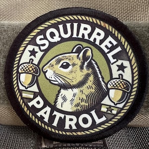 Könnte beinhalten: Ein schwarz-weißes Patch mit einem grün-braun-weißen Tarnmuster. Das Patch zeigt ein Cartoon-Eichhörnchen mit dem Text "Squirrel Patrol" in einem weißen Kreis.