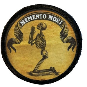 May include: A round, black-edged patch with a yellow background. The patch features a black skeleton kneeling in prayer with wings on either side. The text "MEMENTO MORI" is written in a white banner above the skeleton.