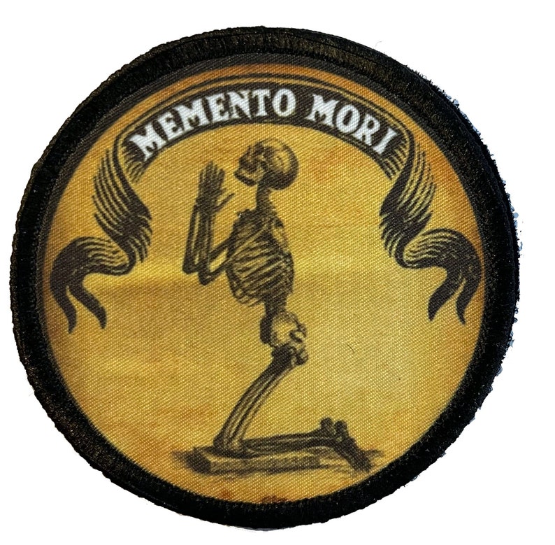May include: A round, black-edged patch with a yellow background. The patch features a black skeleton kneeling in prayer with wings on either side. The text "MEMENTO MORI" is written in a white banner above the skeleton.
