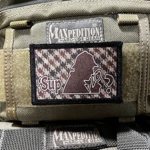 May include: A brown and black camouflage patch with the text "Sup?" and a silhouette of a person wearing a hat and holding a magnifying glass. The patch is attached to a green Maxpedition bag with the text "Hard-Use Gear".
