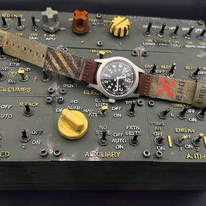 May include: A wristwatch with a black analog face and white numerals, featuring a canvas strap with brown leather accents, black and yellow hazard stripes, and a red cross symbol, resting on a dark green aircraft control panel with toggle switches.