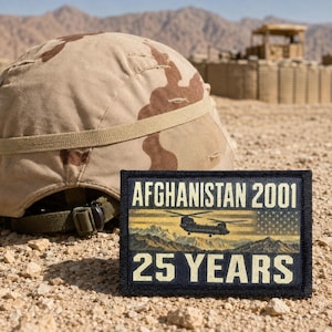 May include: A rectangular patch with a black border featuring the text AFGHANISTAN 2001 and 25 YEARS in beige. The center displays a graphic of a helicopter flying over mountains with a faded American flag overlay, resting near a tan camouflage helmet.