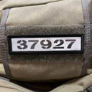 Könnte beinhalten: Eine graue taktische Tasche mit einem schwarzen Patch, auf dem "37927" steht.