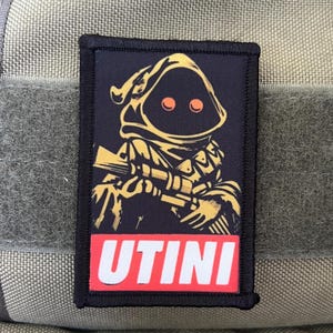 May include: A black and yellow embroidered patch with a cartoon character holding a weapon. The character has a hooded robe and red eyes. The text "UTINI" is printed in red on a white background.