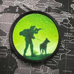 May include: A green and black embroidered patch featuring a silhouette of a person with a rifle and a dog. The patch is circular and has a black border.