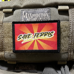 May include: A black and red patch with a yellow sunburst design and the text "Save Ferris" in a yellow, handwritten font. The patch is attached to a green tactical bag with the Maxpedition Hard-Use Gear logo.