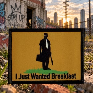 May include: A rectangular yellow patch with a black border featuring a black silhouette of a person in a short-sleeved shirt and tie holding a briefcase and a shotgun, standing on green grass with the text I Just Wanted Breakfast below.