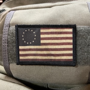 May include: A gray backpack with a black patch featuring a vintage-style American flag with 13 stars in a circle on the blue field.
