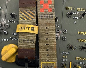 May include: Two olive green canvas watch straps with brown leather accents. The left strap features black and yellow diagonal stripes, a yellow loop labeled UNIT 4, and the word CREW. The right strap has a red cross, the text ENG, and a yellow box with EVA AUTH.