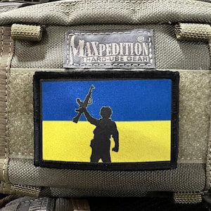 May include: A black patch with a yellow and blue Ukrainian flag and a silhouette of a person holding a rifle. The text "Maxpedition Hard-Use Gear" is visible on the bag.