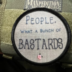 May include: A black and white embroidered patch with the text "PEOPLE. WHAT A BUNCH OF BASTARDS" on a grey background. The patch is attached to a green bag with a black Maxpedition Hard Use Gear label.