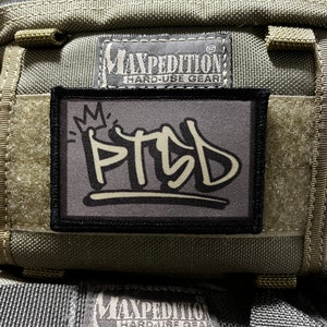 Op de afbeelding: Een groene Maxpedition Hard-Use Gear tas met een zwarte patch waarop "PTSD" staat in witte graffiti-letters met een kroon boven de tekst.