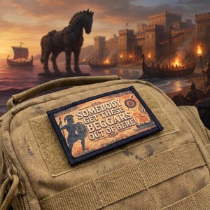 Puede incluir: Un parche rectangular con borde negro fijado en una bolsa táctica de color tostado. El parche presenta un fondo naranja y marrón con una ilustración de un guerrero y el texto: SOMEBODY GET THESE BEGGARS OUT OF HERE!