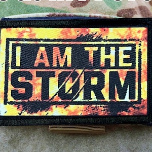 May include: A black and yellow patch with the words "I AM THE STORM" in white letters on a fiery orange background.