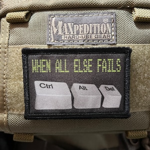Könnte beinhalten: Ein schwarzer Patch mit weißem Text, der "WHEN ALL ELSE FAILS" lautet, und drei weiße Computertasten mit dem Text "Ctrl", "Alt" und "Del". Der Patch ist an einem grünen taktischen Rucksack mit einem schwarzen Etikett "MAXPEDITION HARD-USE GEAR" befestigt.