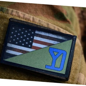 May include: Rectangular patch featuring the American flag on the top left and a green triangle with a blue design on the bottom right. The patch is attached to a camouflage fabric.