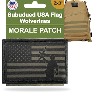 Könnte beinhalten: Ein schwarzer und grüner Morale-Patch mit einem gedämpften amerikanischen Flaggen-Design und einer Silhouette einer Person, die ein Gewehr hält. Der Patch ist ca. 5 x 7,6 cm groß und trägt die Aufschrift "Subdued USA Flag Wolverines Morale Patch". Ein beigefarbener Rucksack ist im Hintergrund.