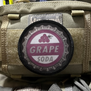 Puede incluir: Un parche blanco y negro con un diseño de tapón de botella de soda de uva púrpura. El parche tiene las palabras "Grape Soda" en texto blanco sobre un fondo púrpura.