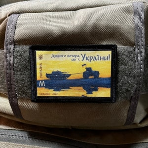 May include: A black and yellow patch with the Ukrainian flag and the text "Доброго вечора, Україна!" (Good evening, Ukraine!) and a depiction of a boat and a tractor. The patch is labelled "Україна 2022" (Ukraine 2022).
