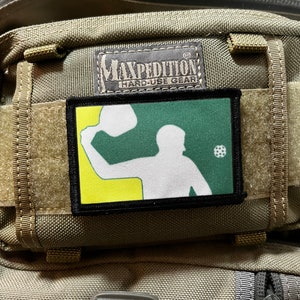 Puede incluir: Un parche verde, amarillo y blanco con una silueta de una persona jugando un deporte con una pala y una pelota. El parche está unido a una mochila táctica marrón con el texto "Maxpedition Hard-Use Gear" en una etiqueta negra.