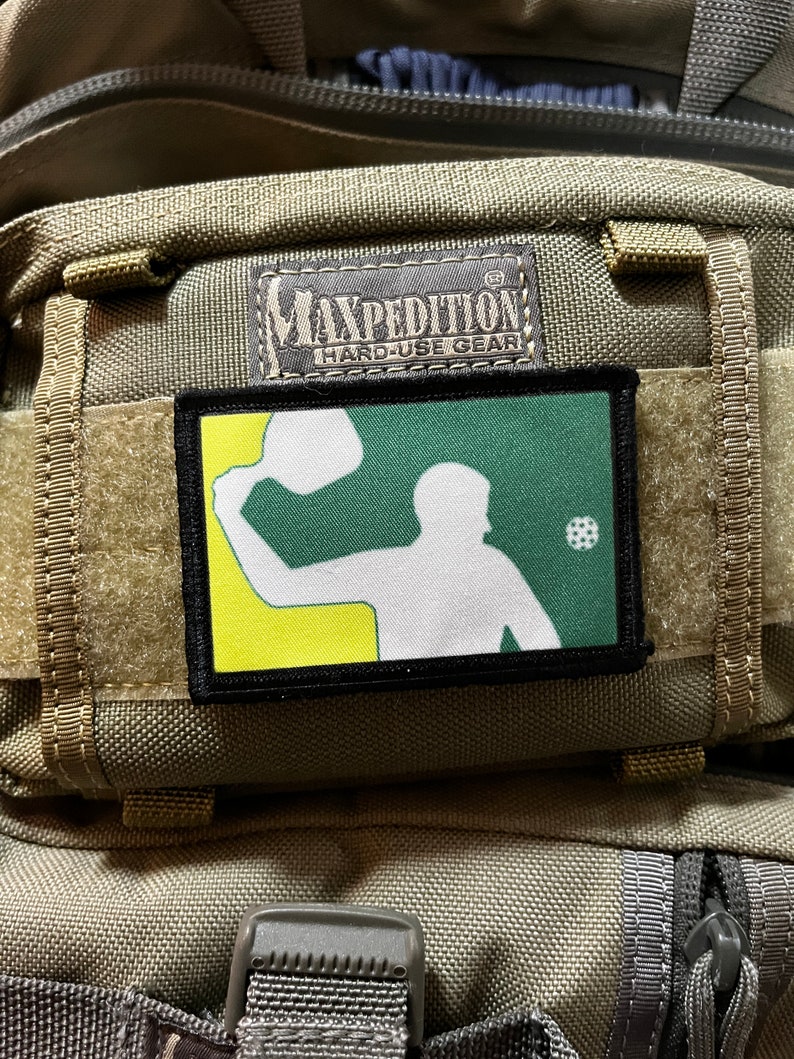 Pu&ograve; includere: Una toppa verde, gialla e bianca con la silhouette di una persona che pratica uno sport con una racchetta e una palla. La toppa &egrave; attaccata a uno zaino tattico marrone con il testo "Maxpedition Hard-Use Gear" su un'etichetta nera.