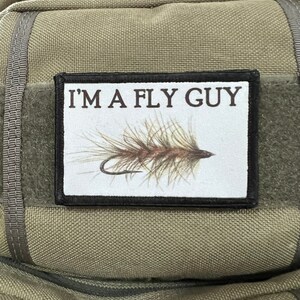 May include: A rectangular patch with the text "I'M A FLY GUY" above a detailed illustration of a fishing fly. The fly is brown and tan with a curved hook. The patch is on a green fabric background.