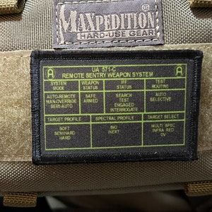 Könnte beinhalten: Ein schwarzes und grünes Etikett mit dem Text "UA 571-C REMOTE SENTRY WEAPON SYSTEM" und einer Tabelle mit den Überschriften "SYSTEM MODE", "WEAPON STATUS", "RF STATUS", "TEST ROUTINE". Die Tabelle listet verschiedene Einstellungen für das System auf.