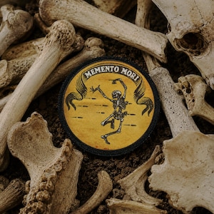Könnte beinhalten: Ein runder Aufnäher mit der Aufschrift "Memento Mori" und einem tanzenden Skelett auf gelbem Grund. Der Aufnäher ist von einem schwarzen Rand umgeben und vor dem Hintergrund von Knochen und Erde platziert.
