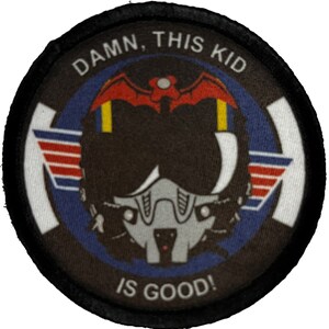May include: A black and white round patch with a black border. The patch has a white background with a blue circle in the center. Inside the blue circle is a black and white illustration of a pilot's helmet with a red bat on top. The text "DAMN, THIS KID IS GOOD!" is written in white around the blue circle.