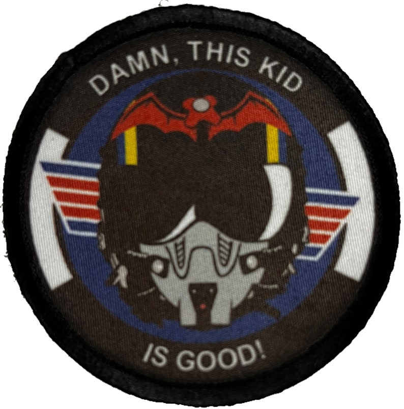 May include: A black and white round patch with a black border. The patch has a white background with a blue circle in the center. Inside the blue circle is a black and white illustration of a pilot's helmet with a red bat on top. The text "DAMN, THIS KID IS GOOD!" is written in white around the blue circle.