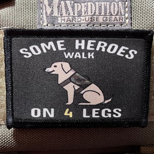 May include: Black rectangular patch with white text that reads "SOME HEROES WALK ON 4 LEGS." A tan dog wearing a vest is in the center. The patch is on a green and tan background.