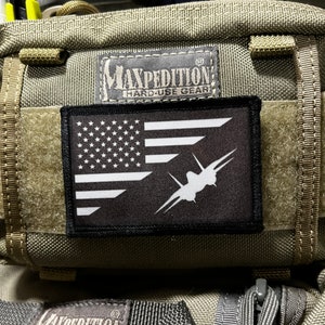 Puede incluir: Un parche negro con una bandera estadounidense blanca y una silueta blanca de un avión de combate. El parche está unido a una bolsa táctica verde con el texto "Maxpedition Hard-Use Gear" en ella.