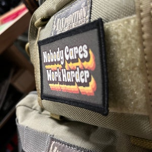 May include: A close-up of a Maxpedition Hard Use Gear bag with a black patch that reads "Nobody Cares Work Harder" in a retro font with a flame design. The patch is attached to the front of the bag.