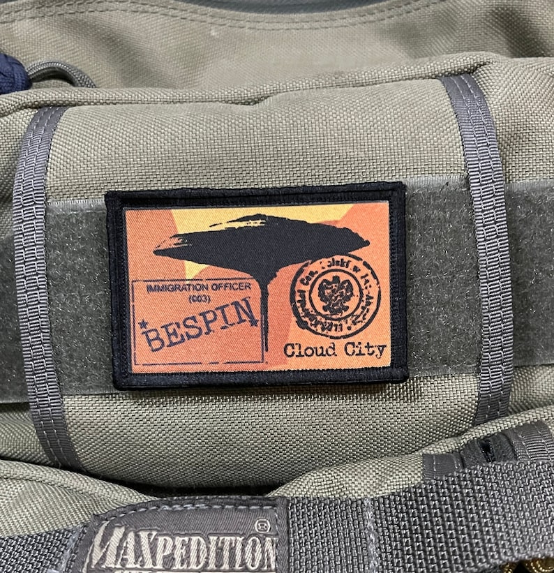 May include: A black and orange patch with the text "Immigration Officer (CO3) Bespin Cloud City" and a graphic of a flying saucer. The patch is attached to a green bag.