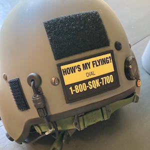 May include: A green military helmet with a black velcro patch and a yellow sticker that reads "How's My Flying? Dial 1-800-SQK-7700".